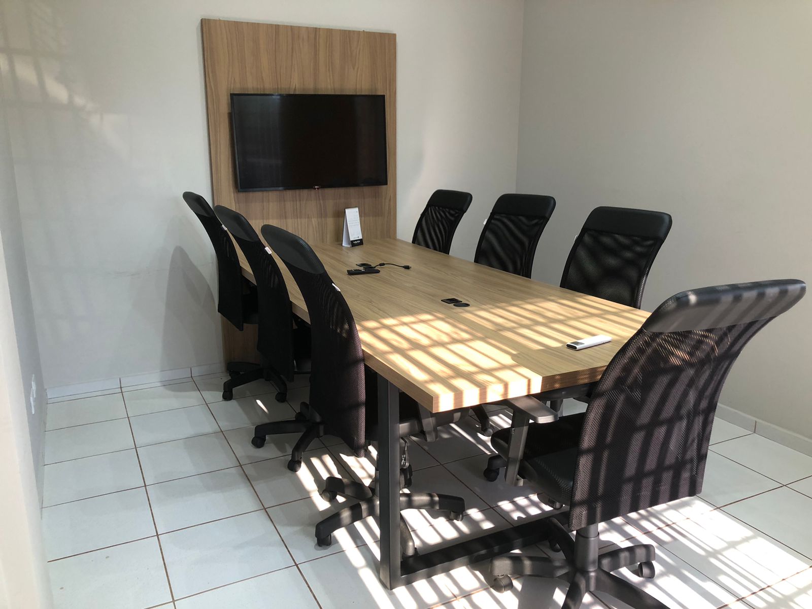 Sala de reunião iluminada com luz natural, mesa grande e TV na parede
