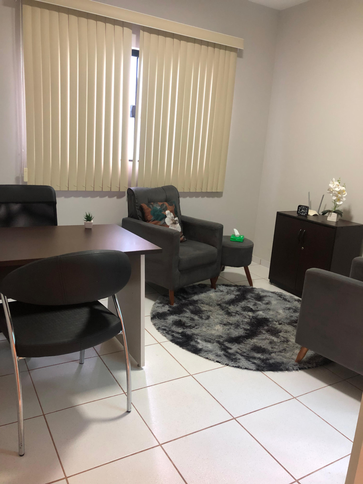 Consultório com mesa de trabalho, poltronas cinza e tapete redondo